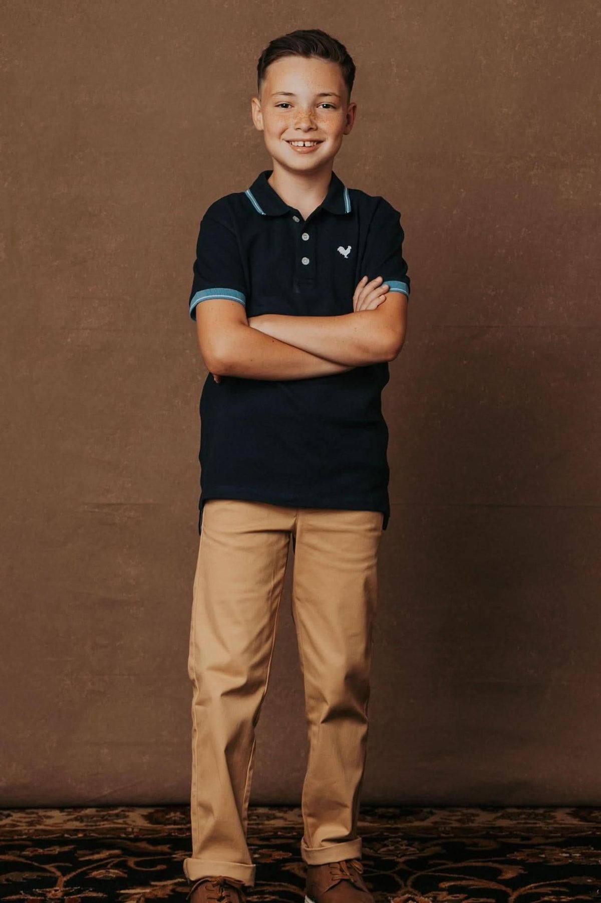 A smiling boy stands confidently with crossed arms, wearing a navy polo shirt and khaki chinos against a brown backdrop.