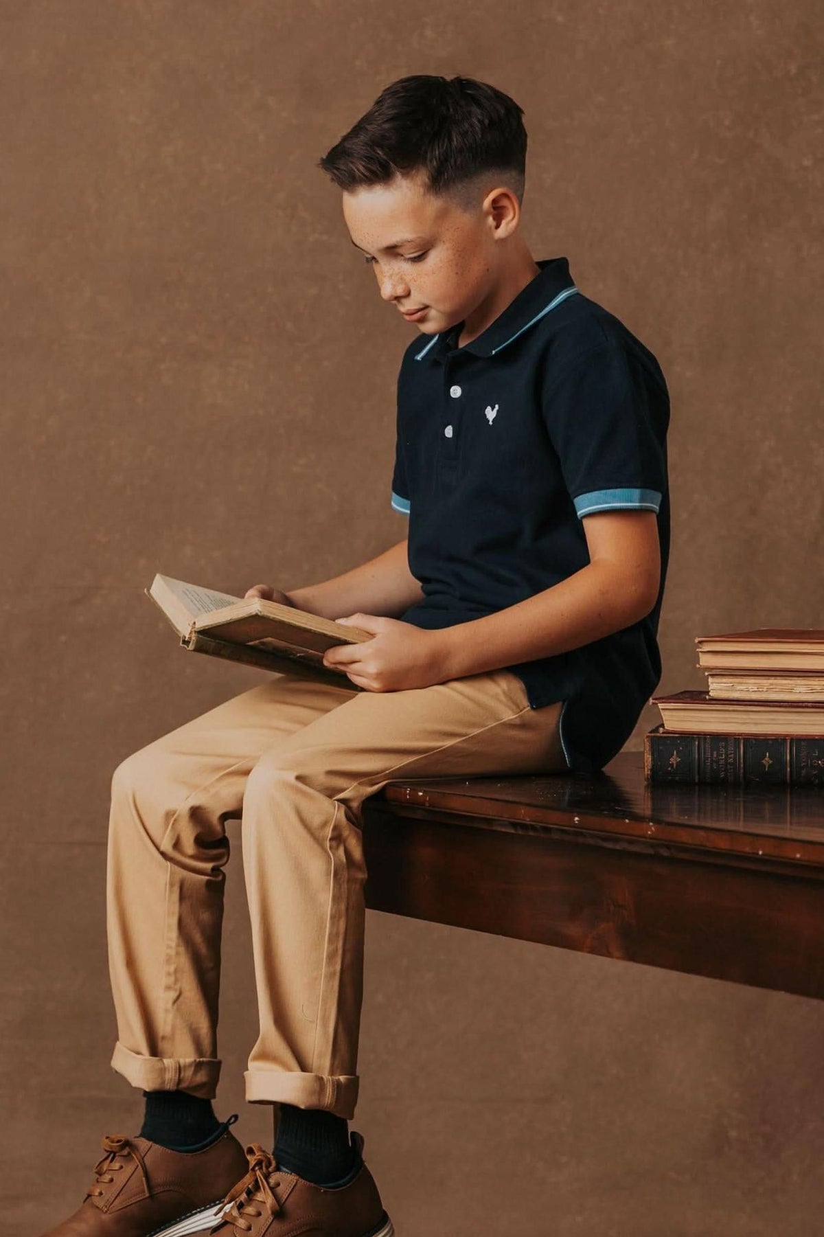 A boy in a navy polo and khaki chinos sits on a wooden table, reading a book with vintage-style books.