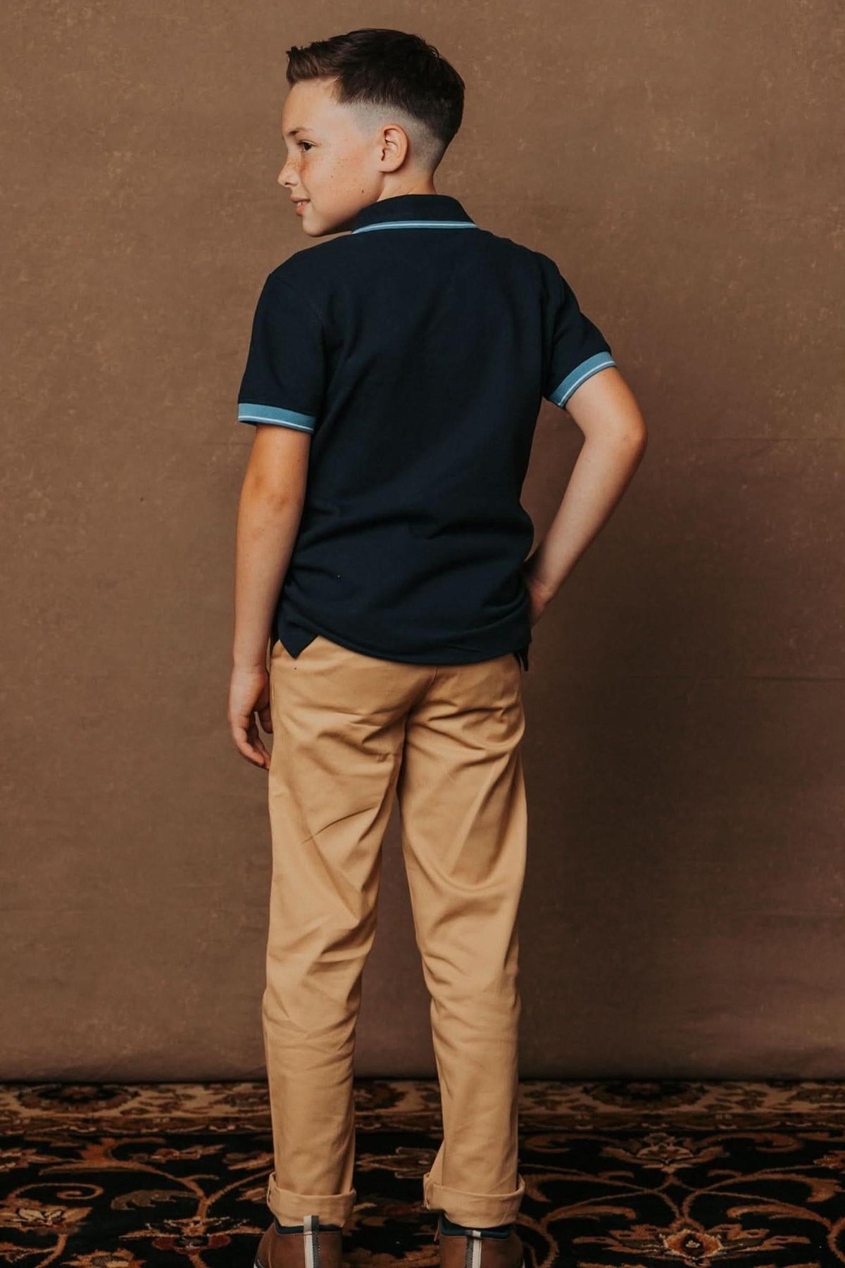 A boy in a navy polo shirt and khaki chinos poses with his back to the camera against a textured.