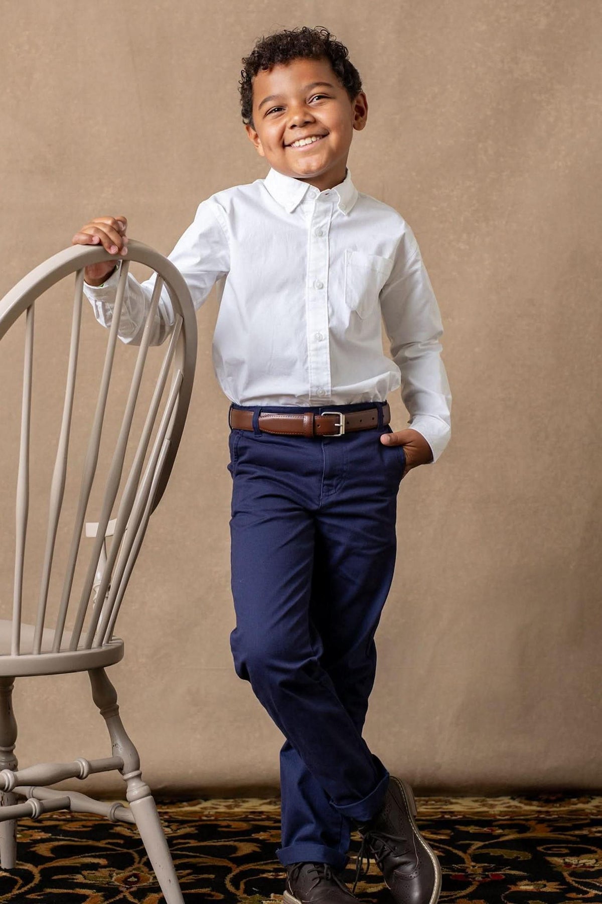 A smiling boy stands next to a gray chair, wearing a white shirt, navy chino pants, and dark shoes.