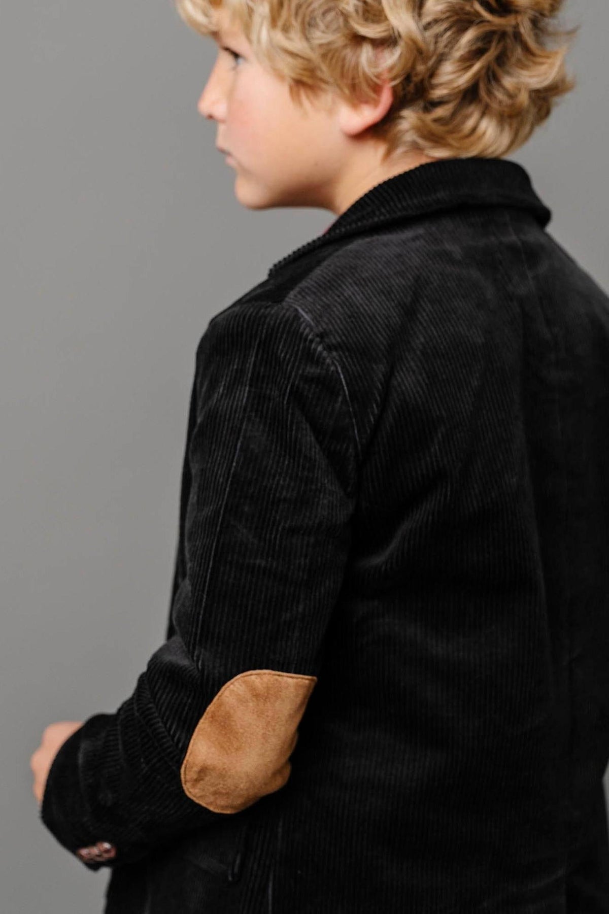 A young boy models a black corduroy blazer, featuring faux suede elbow patches. The tailored silhouette emphasizes a classic and stylish look suitable for kids.