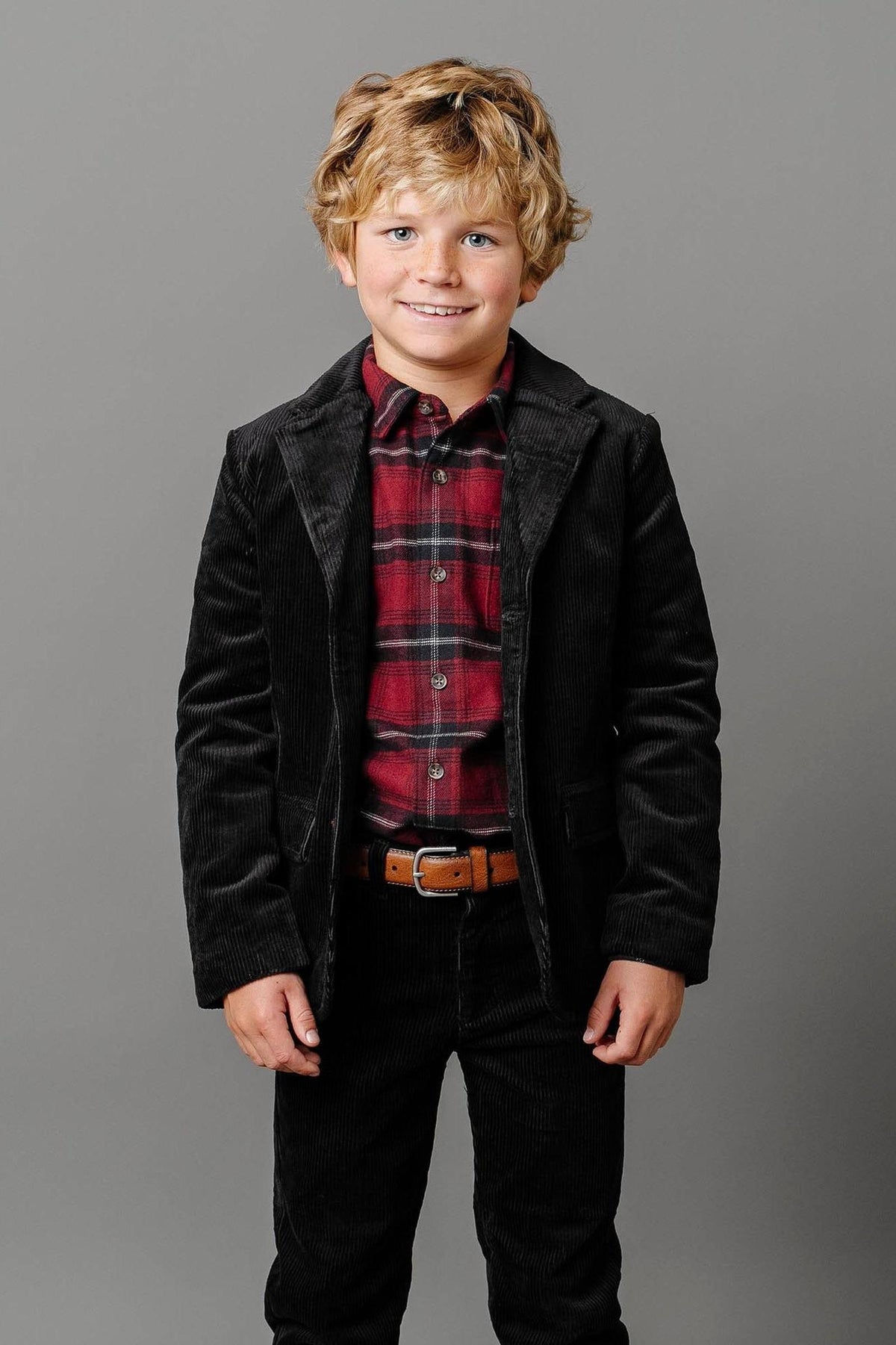 A young boy smiles while wearing a black corduroy blazer with faux suede elbow patches, paired with a red plaid shirt and dark pants, against a gray background.