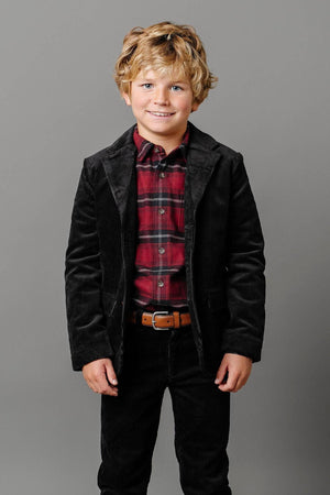 A young boy smiles while wearing a black corduroy blazer with faux suede elbow patches, paired with a red plaid shirt and dark pants, against a gray background.