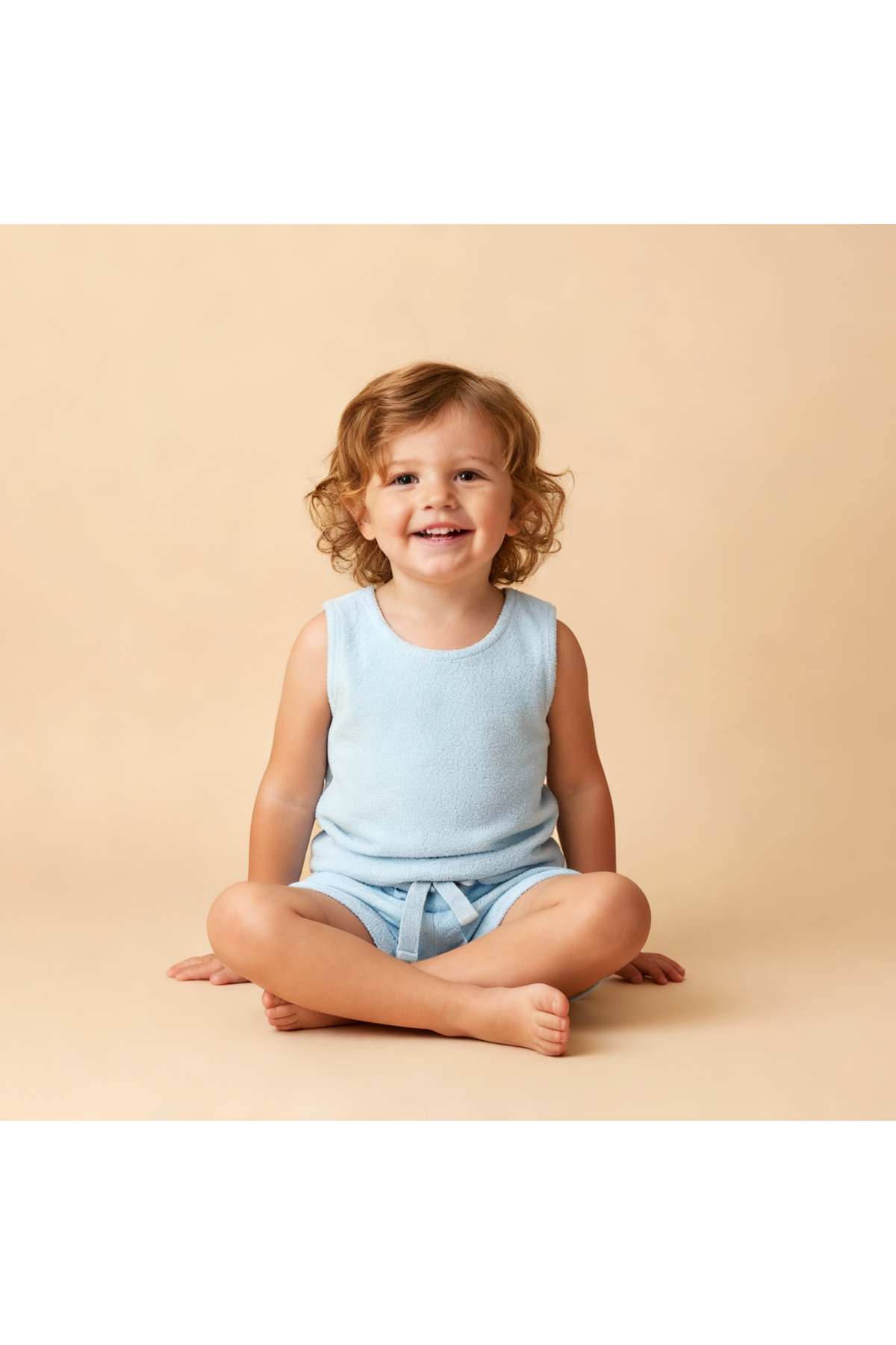 A smiling child sits cross-legged, wearing a light blue organic sleeveless tee and matching shorts set. The soft terry fabric gives a cozy, playful look against a neutral background.