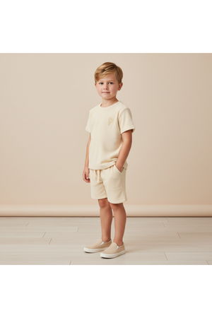 A young boy stands wearing a soft, organic terry butter yellow tee and shorts set, featuring a relaxed fit. The outfit is styled for comfort and summer play.