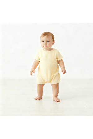 A toddler stands in a light butter yellow organic cotton romper, featuring short sleeves and a relaxed fit. The soft fabric is perfect for warm-weather play and comfort.
