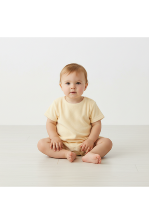 A toddler sits on a light wooden floor, wearing a soft butter yellow organic cotton short romper. The relaxed style is perfect for warm weather and playful activities.
