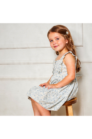 A young girl sits on a wooden stool, wearing a light blue dress with floral patterns and flutter sleeves.