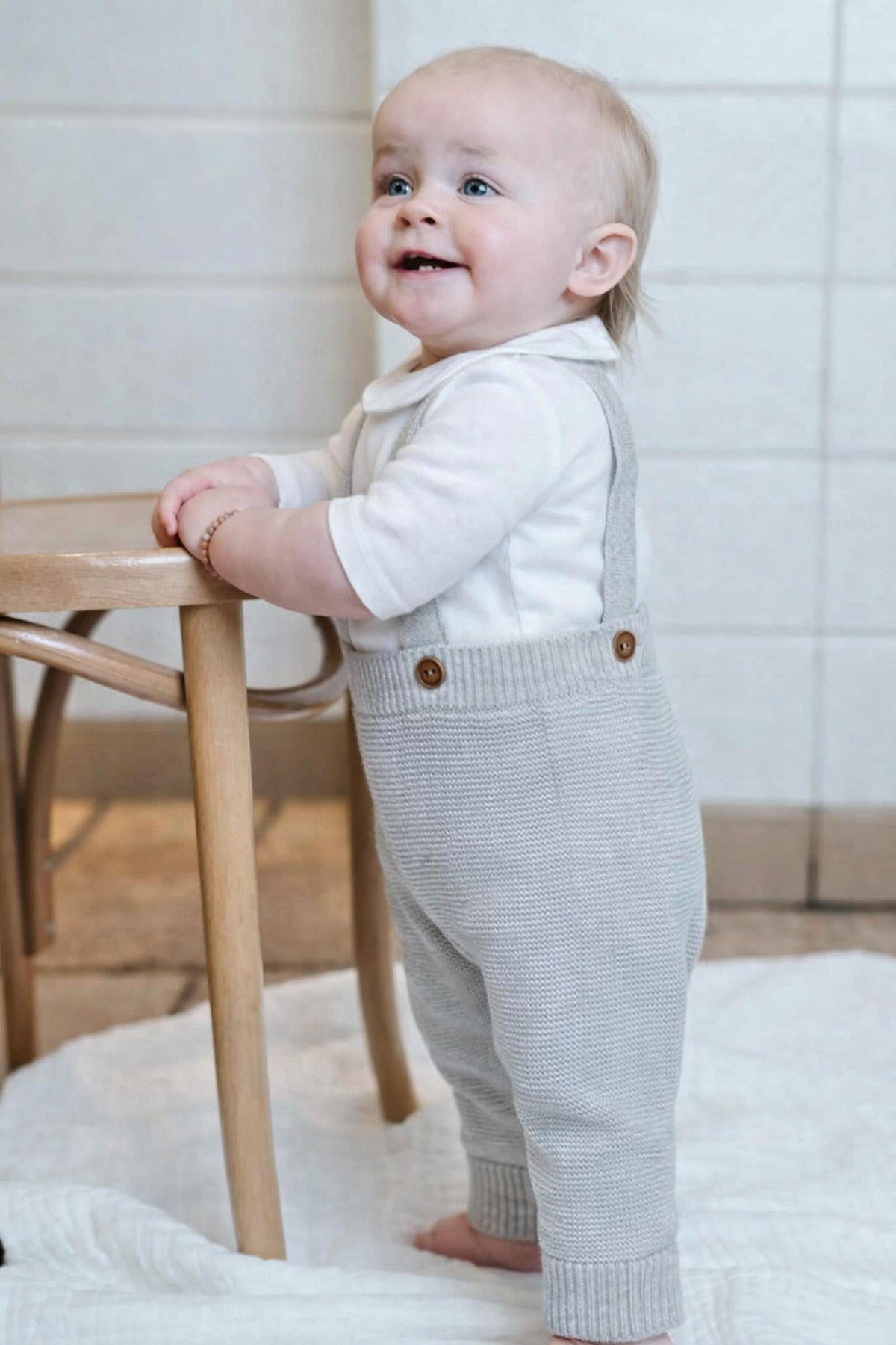 Baby in a knitted grey overalls and white shirt with beige buttons standing on a white rug.
