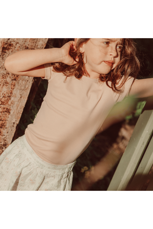 A young girl stands against a wooden background, wearing a light pink fitted top and a floral skirt.