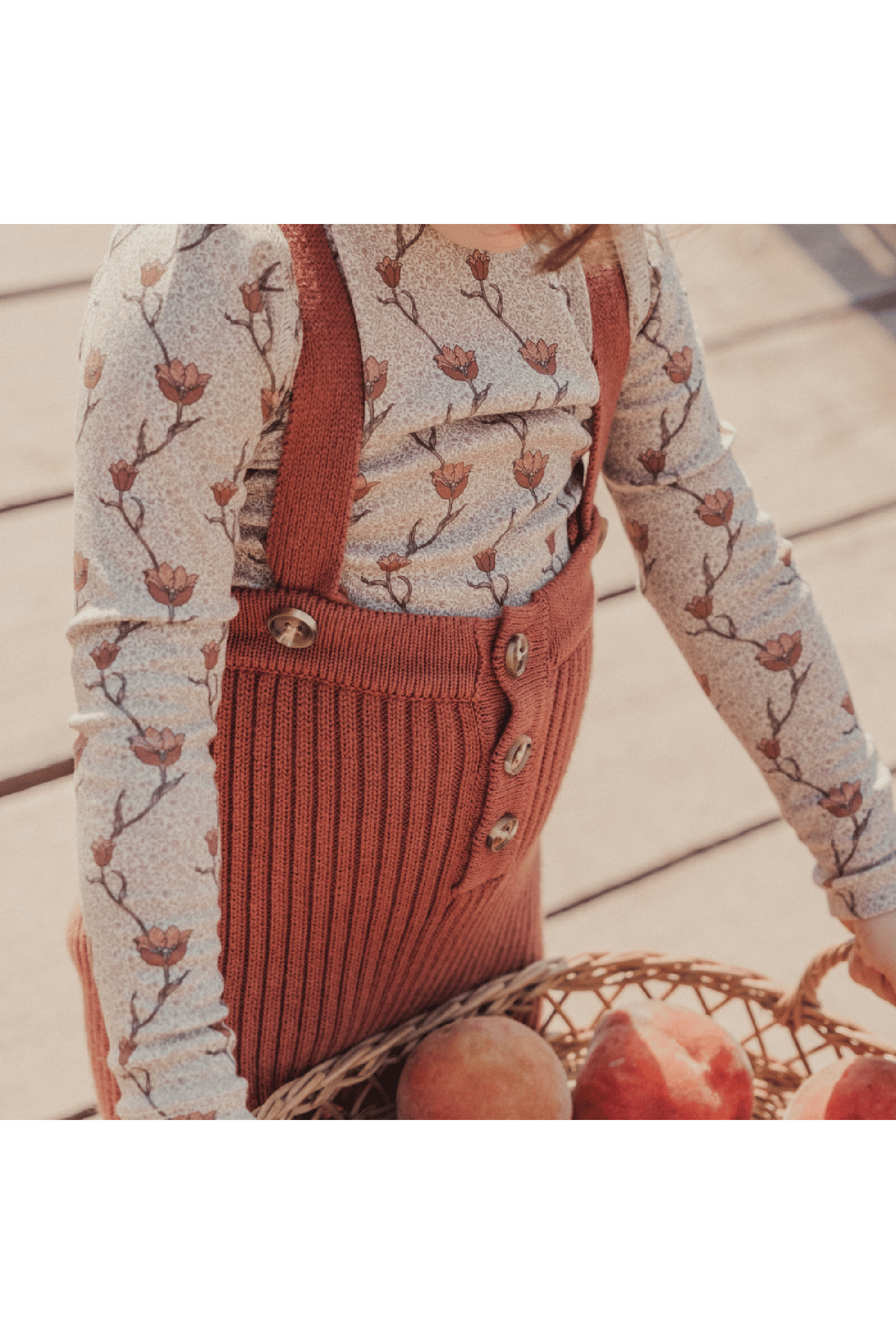 A child wears rosewood knit cotton overalls with a floral long-sleeve top, holding a basket of peaches. The overall features buttons and ribbed detailing, showcasing a cozy, stylish look.