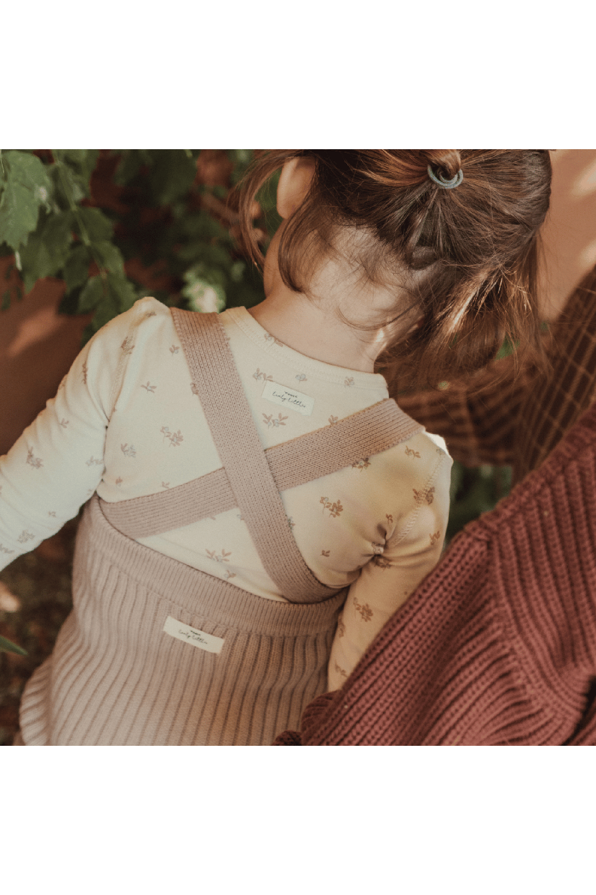 A toddler is wearing a mauve knit overall over a light floral long-sleeve shirt, showcasing the soft texture and cross-back design. The setting features greenery in the background.