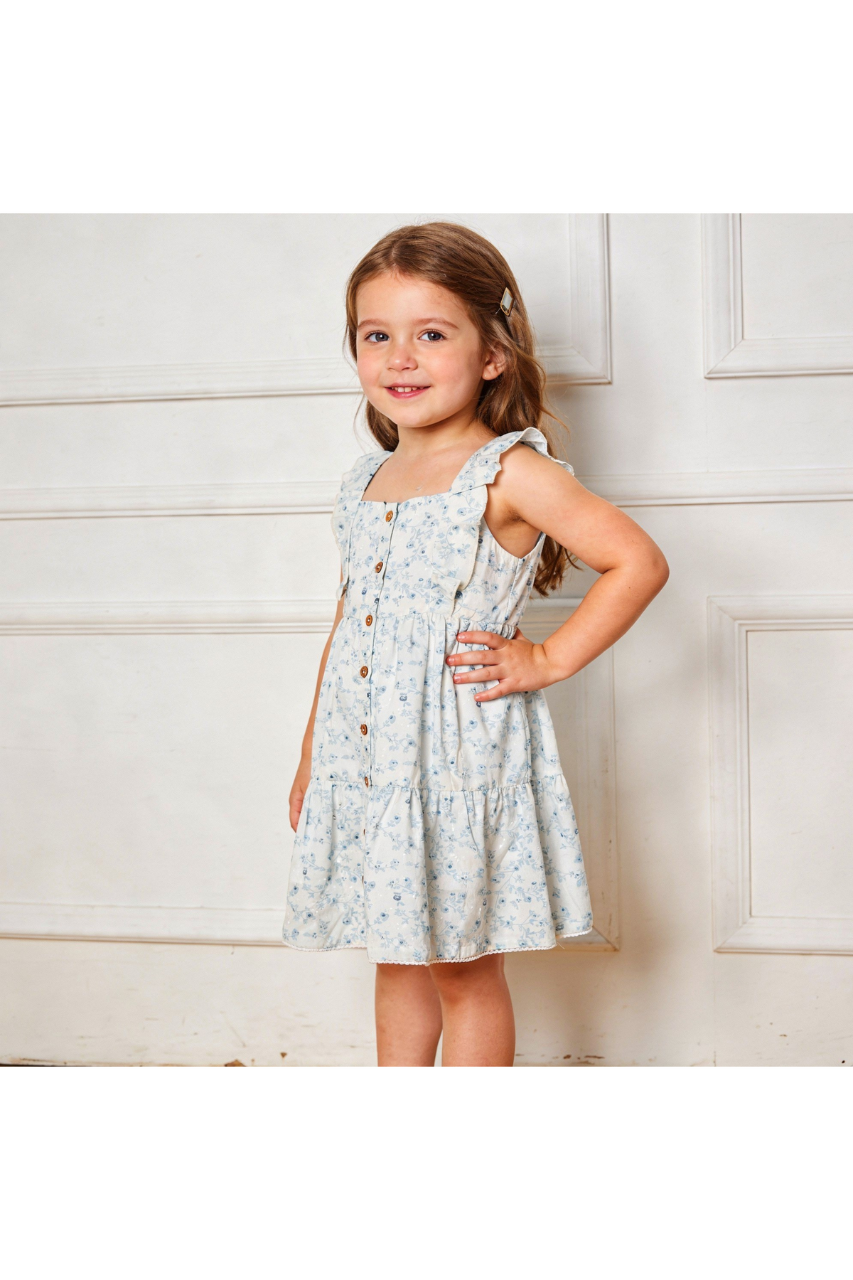 A young girl stands smiling in a light blue, tiered dress with a floral pattern and flutter sleeves. The dress features a button front, showcasing its playful and stylish design.