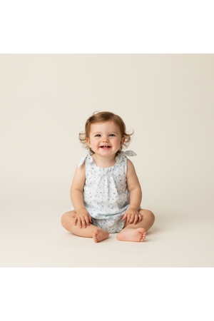 A smiling baby sits on a light background, wearing a soft, floral-patterned romper with tie shoulder straps. The outfit is made from gentle, breathable organic cotton, perfect for comfort.