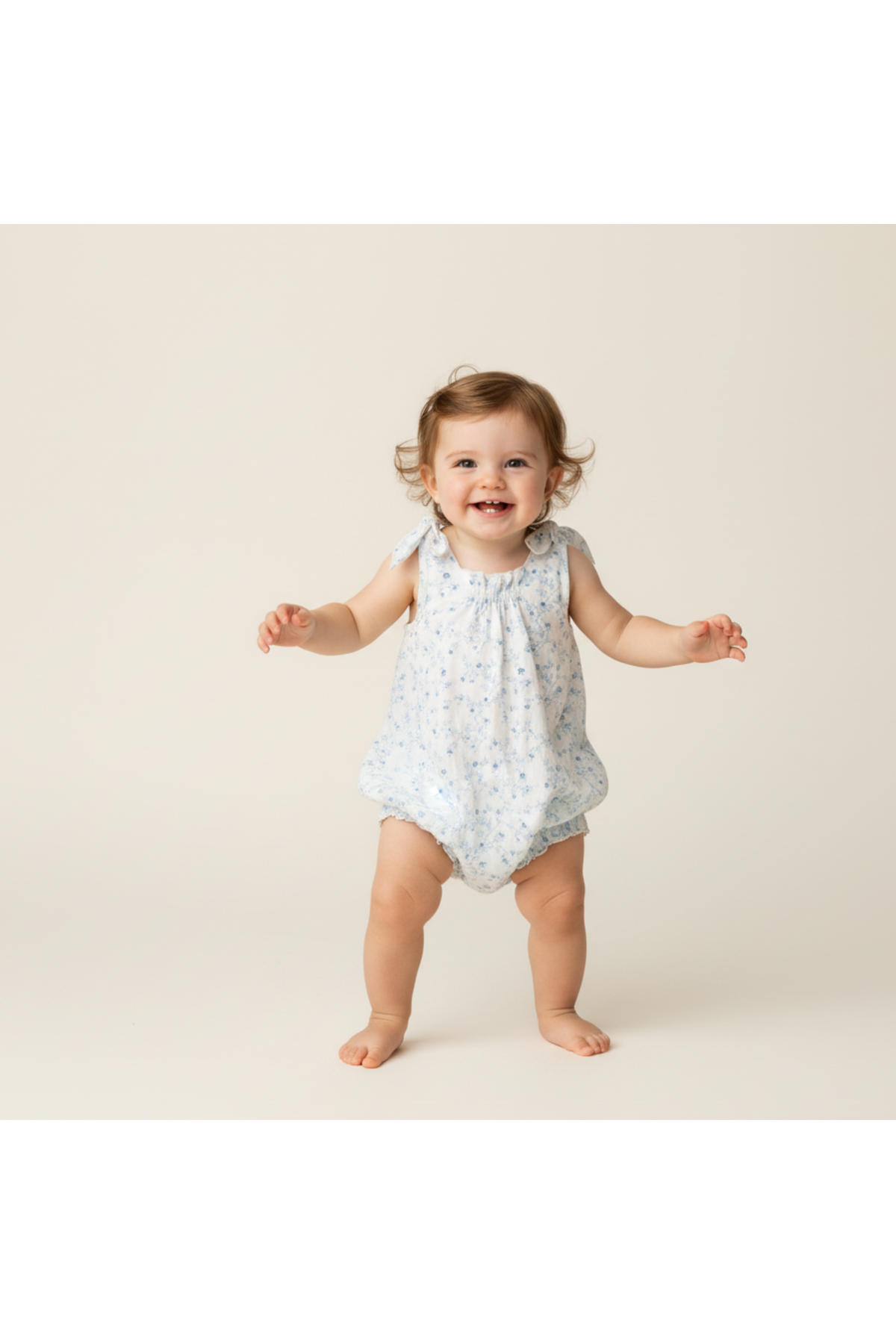 A cheerful toddler wears a light blue bubble romper adorned with tiny floral patterns, featuring tie shoulder straps. The soft fabric is perfect for comfortable, playful movement.