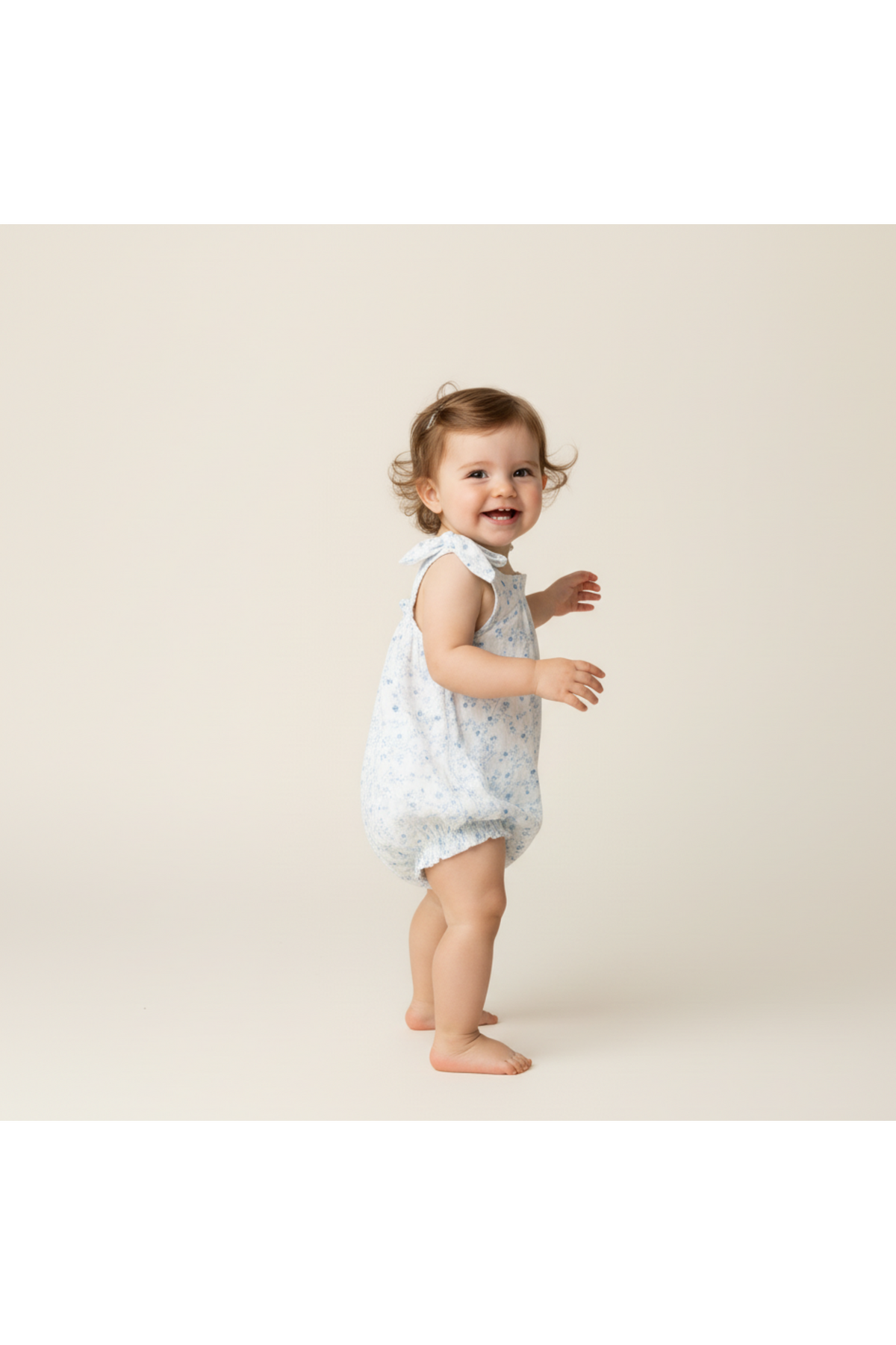 A cheerful toddler with curly hair is wearing a light blue organic cotton bubble romper featuring a delicate floral print.