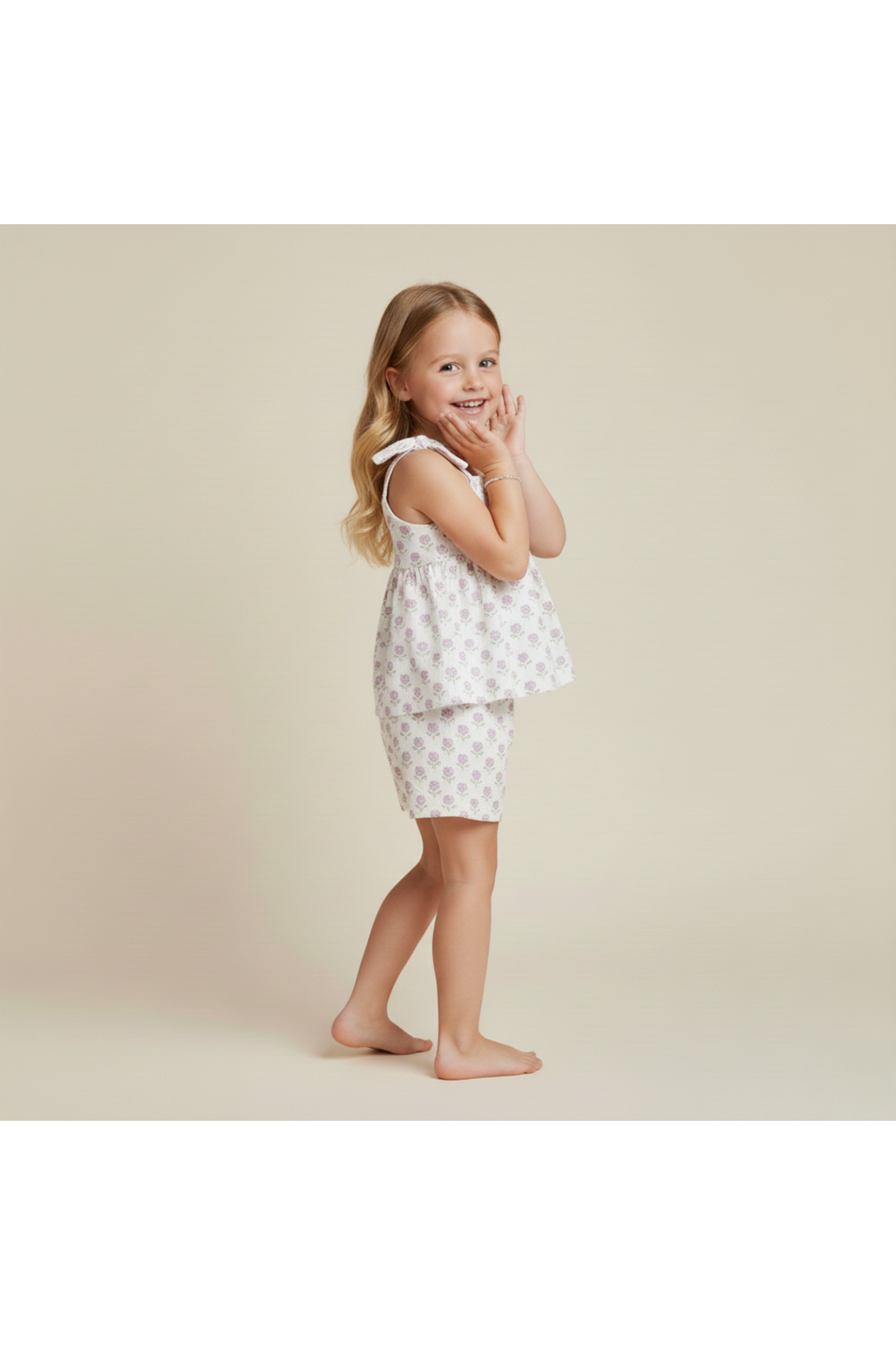 A young girl poses playfully in a lavender and white floral tie-up top and matching shorts set. The outfit features adjustable straps, made from organic cotton, perfect for warm-weather adventures.