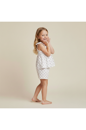A young girl poses playfully in a lavender and white floral tie-up top and matching shorts set. The outfit features adjustable straps, made from organic cotton, perfect for warm-weather adventures.