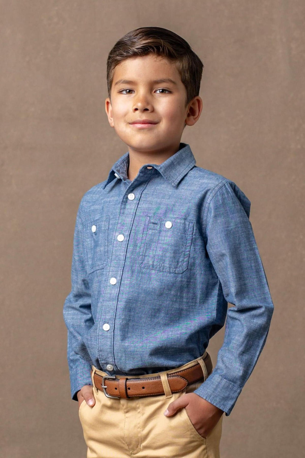 A boy smiles while wearing a blue organic cotton button-down shirt and beige pants, with hands in his pockets.