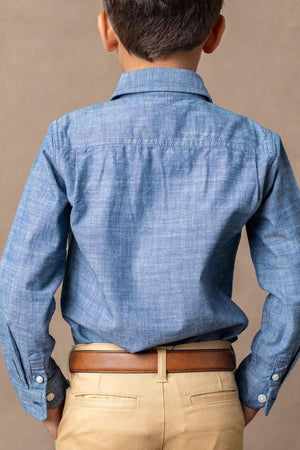 A boy wears a blue organic cotton button-down shirt, paired with khaki pants and a brown belt, against a neutral.