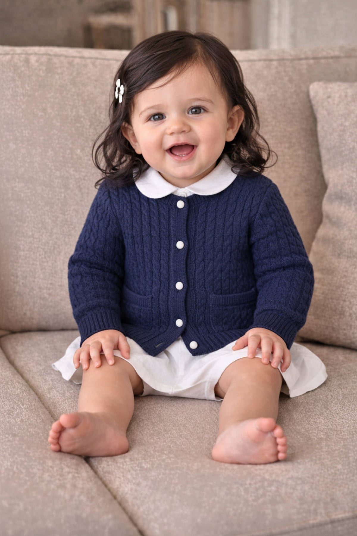 A baby girl sits on a soft beige couch, wearing a navy cable-knit cardigan with white buttons and a collared shirt, paired with a white skirt. She smiles happily.