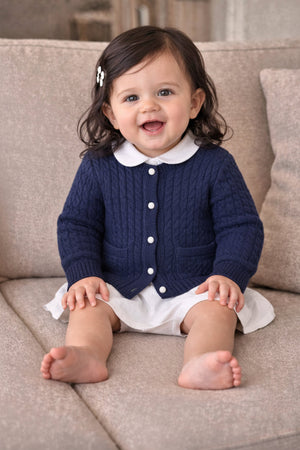 A baby girl sits on a soft beige couch, wearing a navy cable-knit cardigan with white buttons and a collared shirt, paired with a white skirt. She smiles happily.