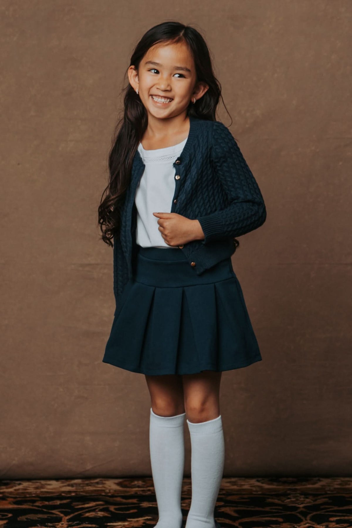 A young girl smiles while wearing a navy cable-knit cardigan over a white shirt and a matching pleated skirt.