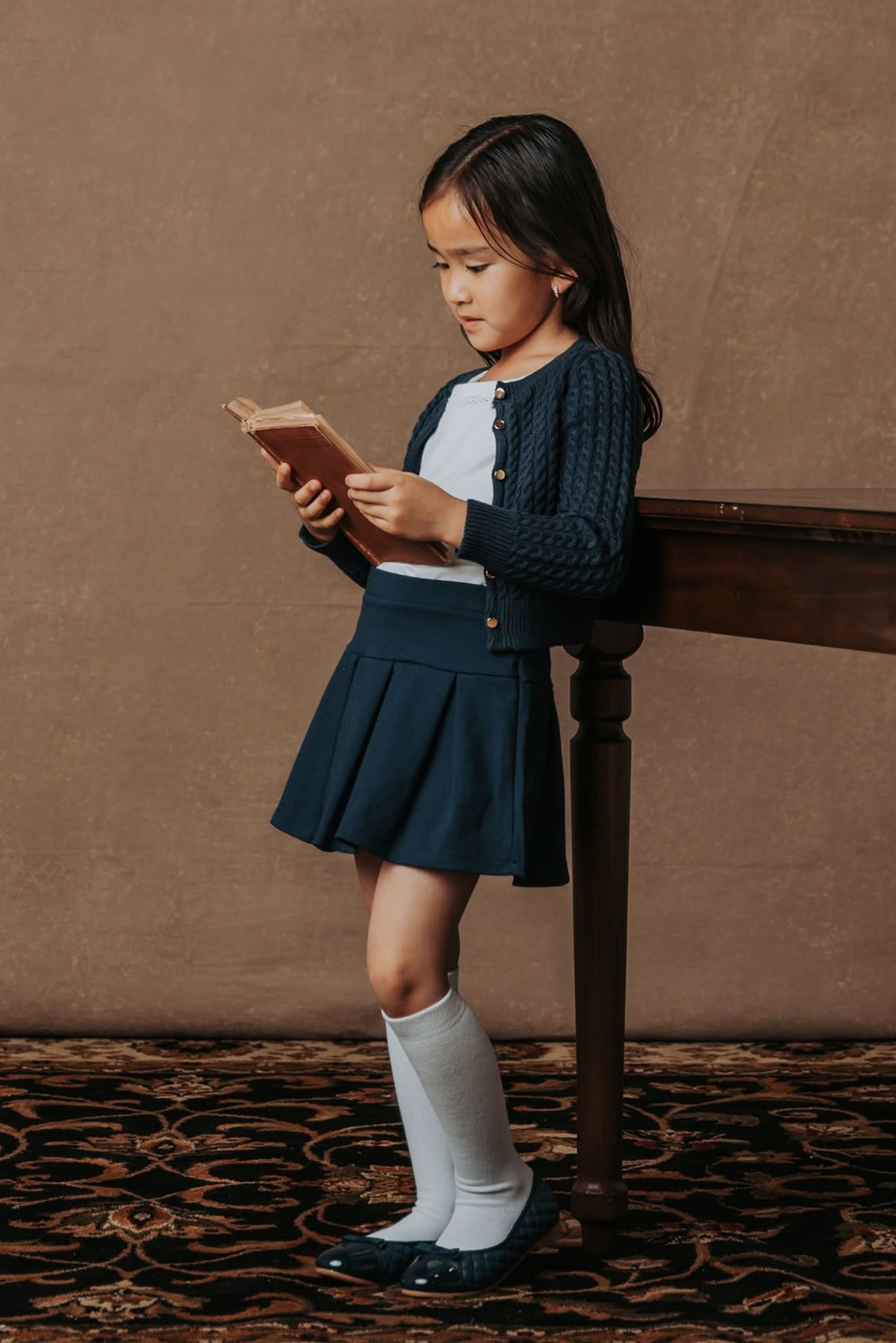 A young girl stands by a wooden table, wearing a navy cable-knit cardigan over a white shirt, paired with a navy pleated skirt and knee-high socks while reading a book.