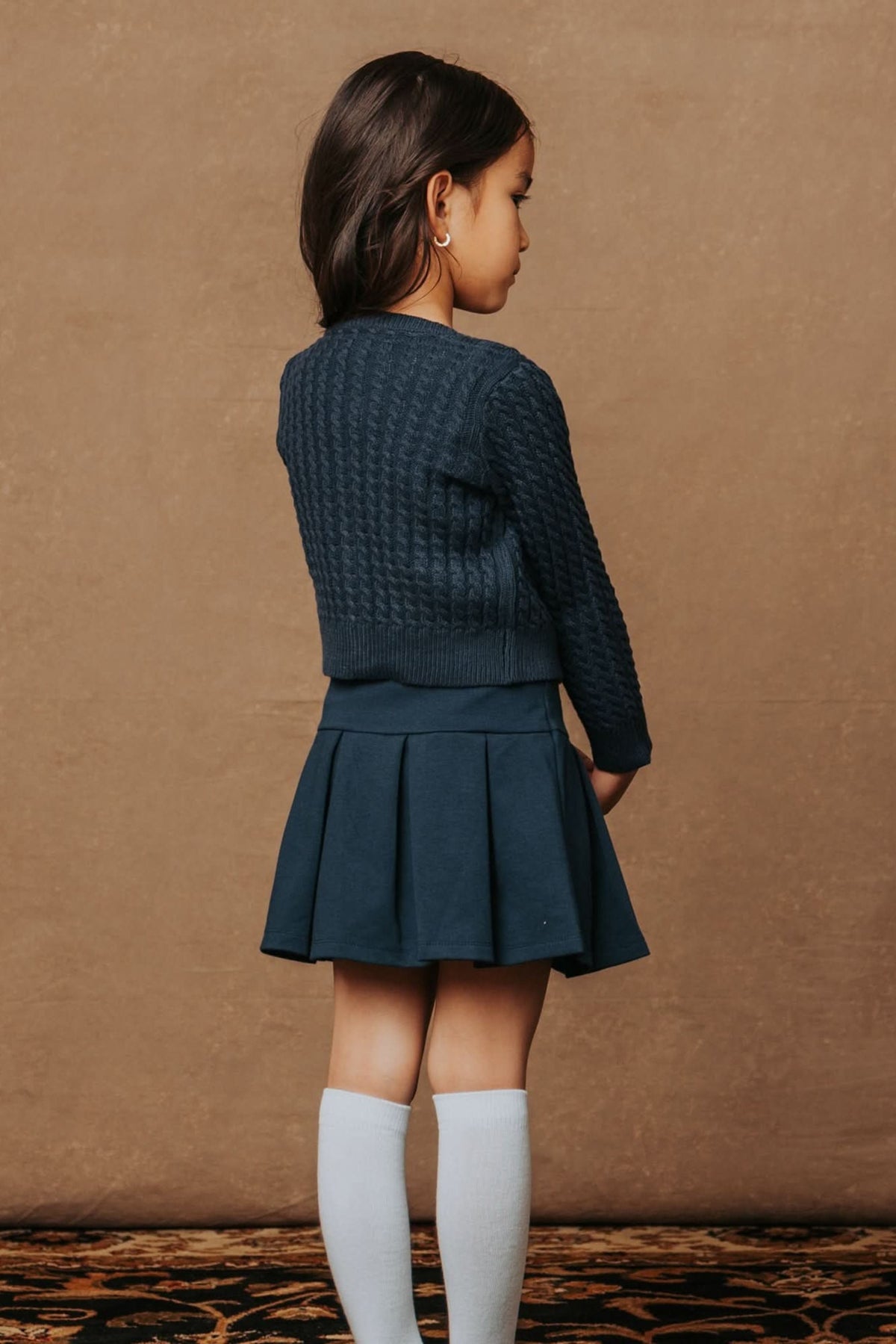 A young girl is seen from the back, wearing a navy cable-knit cardigan and a matching pleated skirt, complemented by knee-high white socks. The background is a warm, neutral tone.