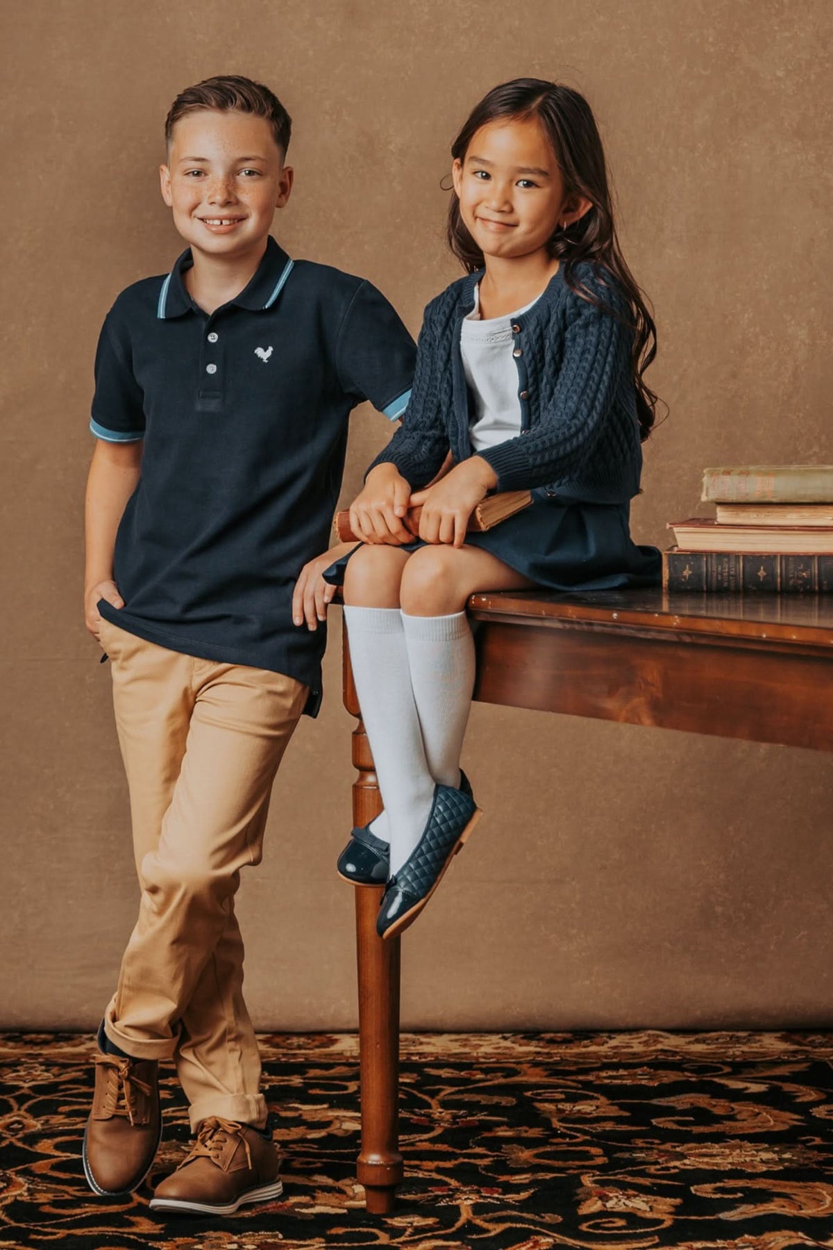 A boy in a navy polo and khaki pants stands beside a girl wearing a navy cable-knit cardigan, white shirt, and skirt, seated on a table with books.
