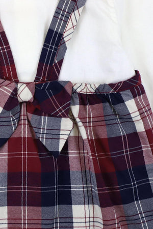 Close-up of a berry tartan plaid jumper featuring a bow and adjustable straps, layered over a white long-sleeve bodysuit with a Peter Pan collar, made from organic cotton.