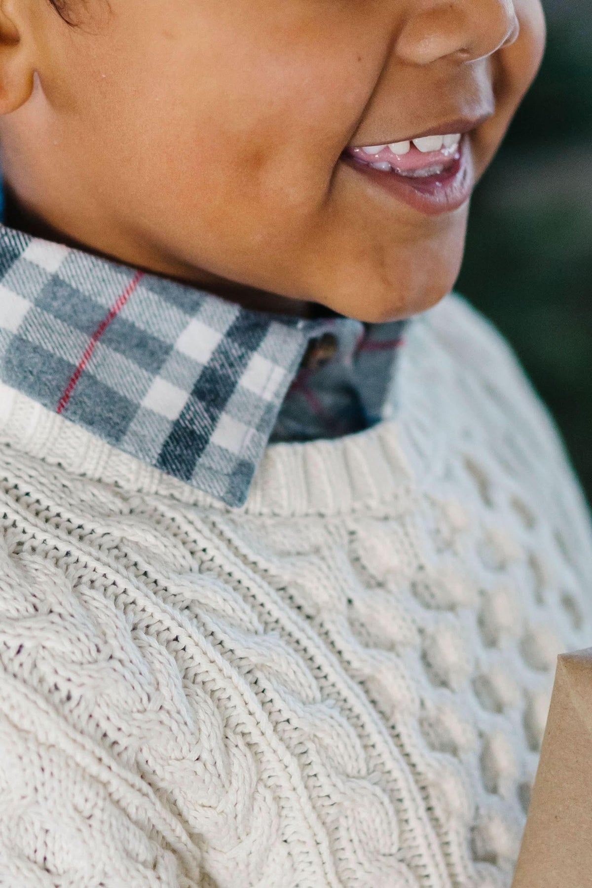 A smiling child wearing a cozy ivory cable knit sweater holds a wrapped gift, complemented by a plaid shirt underneath. The setting features a green backdrop, enhancing the festive atmosphere.