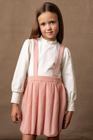 A girl wears a cream long-sleeve top paired with a pleated pink organic cotton overall skirt, set against a neutral.