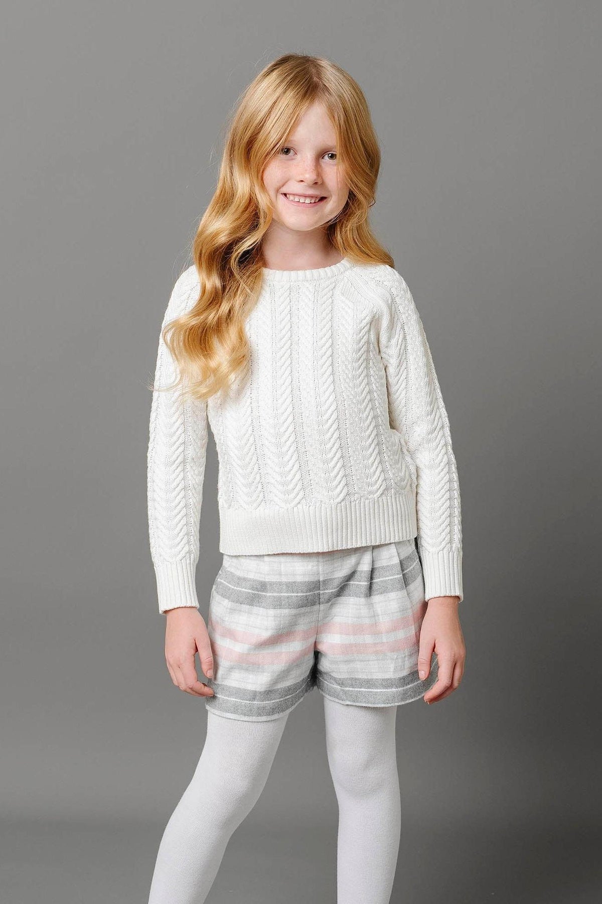 A girl smiles while wearing a cozy white cable-knit sweater paired with plaid pleated shorts in gray and pink, complemented by white tights against a neutral gray background.