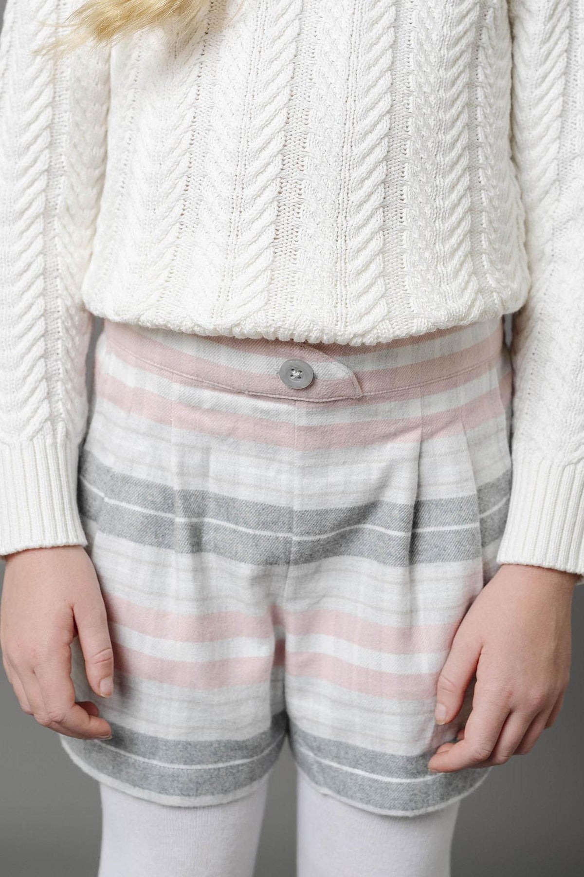 A girl stands with her hands at her sides, wearing soft plaid shorts in pink and gray, paired with a cozy white cable-knit sweater.