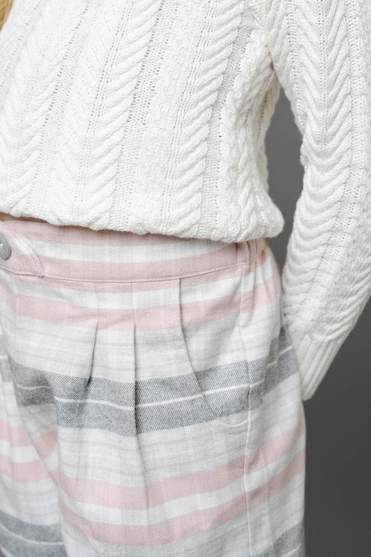 A girl wears a cream cable-knit sweater paired with plaid shorts featuring soft pink, gray, and white stripes. The shorts have a pleated design and a faux button front.