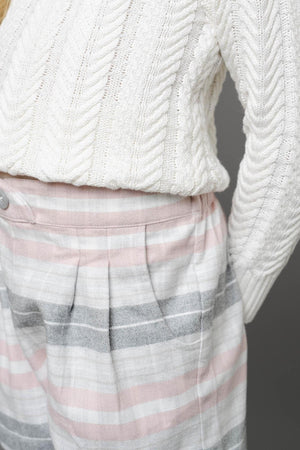 A girl wears a cream cable-knit sweater paired with plaid shorts featuring soft pink, gray, and white stripes. The shorts have a pleated design and a faux button front.