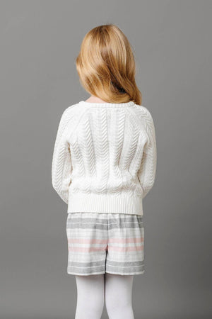 A girl stands with her back to the camera, wearing a cozy white cable-knit sweater and plaid shorts in shades of gray and pink, showcasing a stylish and comfortable outfit.