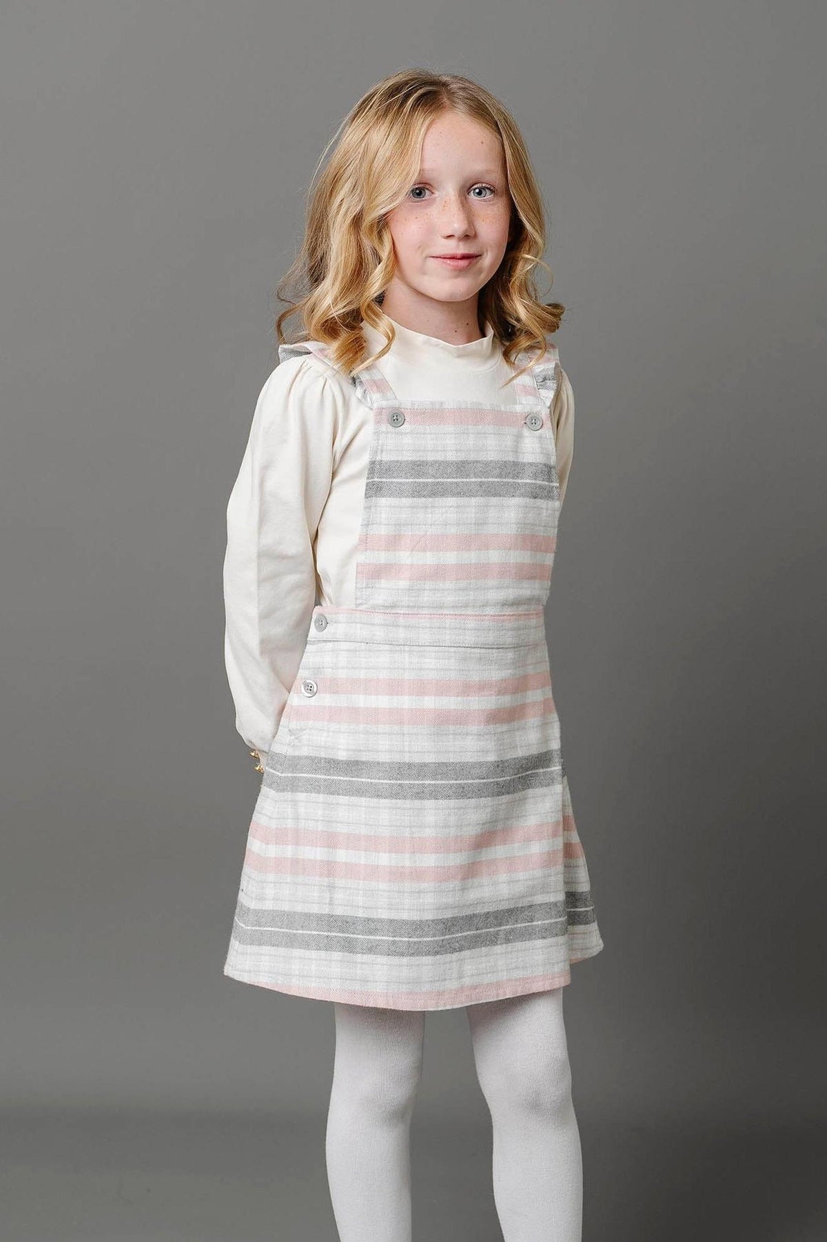 A young girl stands smiling in a gray and rose plaid overall skirt, featuring ruffle straps and button closures.
