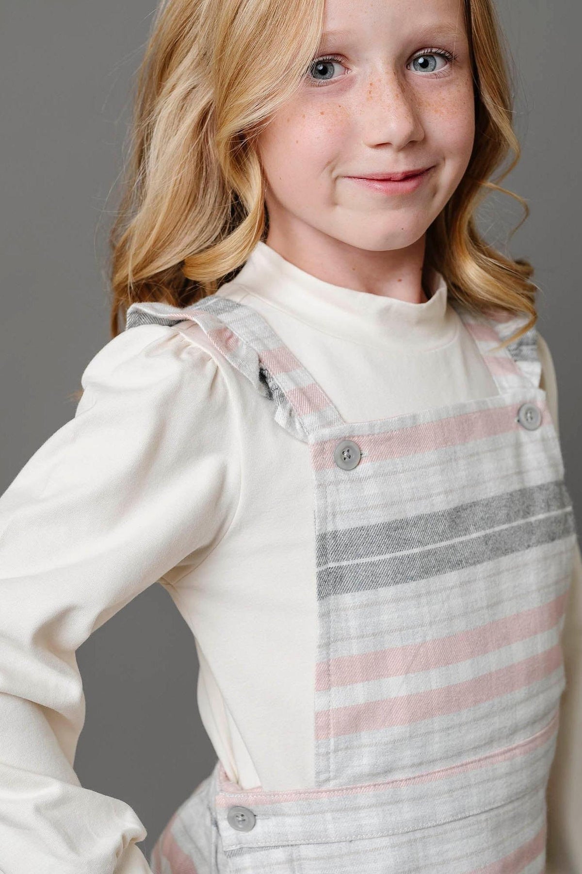 A young girl poses in a gray and rose plaid overall skirt with ruffle straps, made from organic cotton. She wears a cream-colored top, highlighting the skirt’s stylish design.