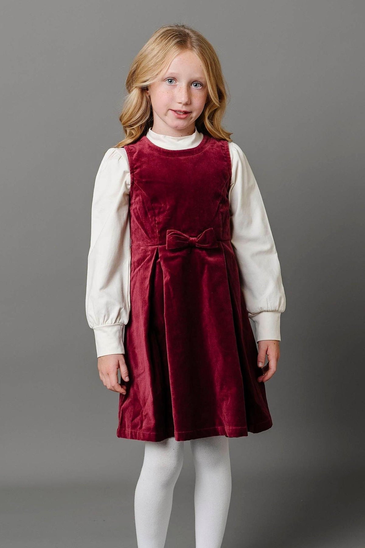 A young girl poses in a wine red velvet jumper dress with a bow front, paired with a white long-sleeve.
