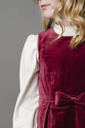 A girl models a wine red jumper dress with a bow front detail, layered over a white top, showcasing soft.