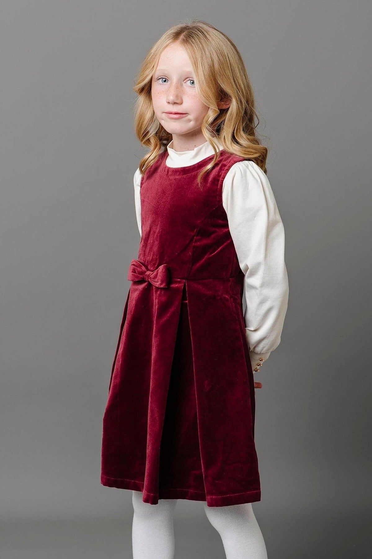 A young girl poses in a wine red velvet jumper dress with a bow front, paired with a white long-sleeve.