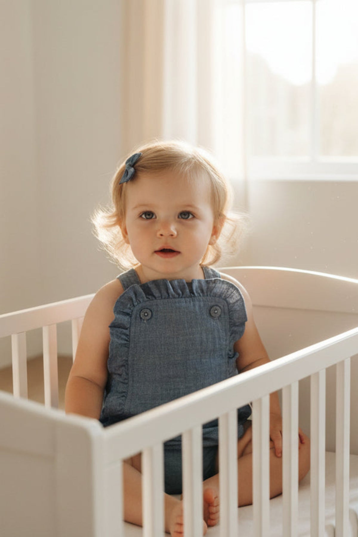 A toddler sits in a white crib, wearing a blue chambray ruffle sunsuit and a matching hair clip.