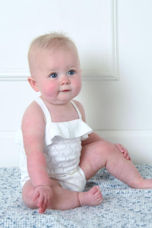 A baby girl with blue eyes sits on a soft, patterned blanket, wearing a white ruffle romper made from organic.