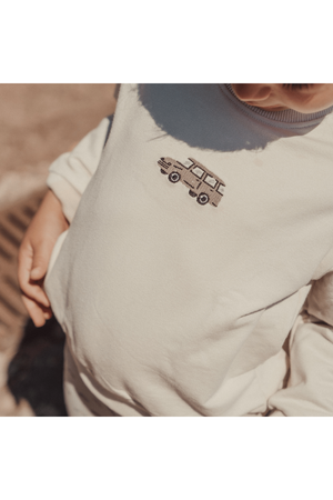 A toddler wearing a cream-colored sweatshirt featuring a vintage car embroidered design. The soft, cotton material is cozy and perfect for casual outings or indoor play.