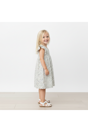 A smiling young girl stands in profile, wearing a light gray organic cotton dress with flutter sleeves and a delicate floral pattern. The background features a simple, neutral setting.