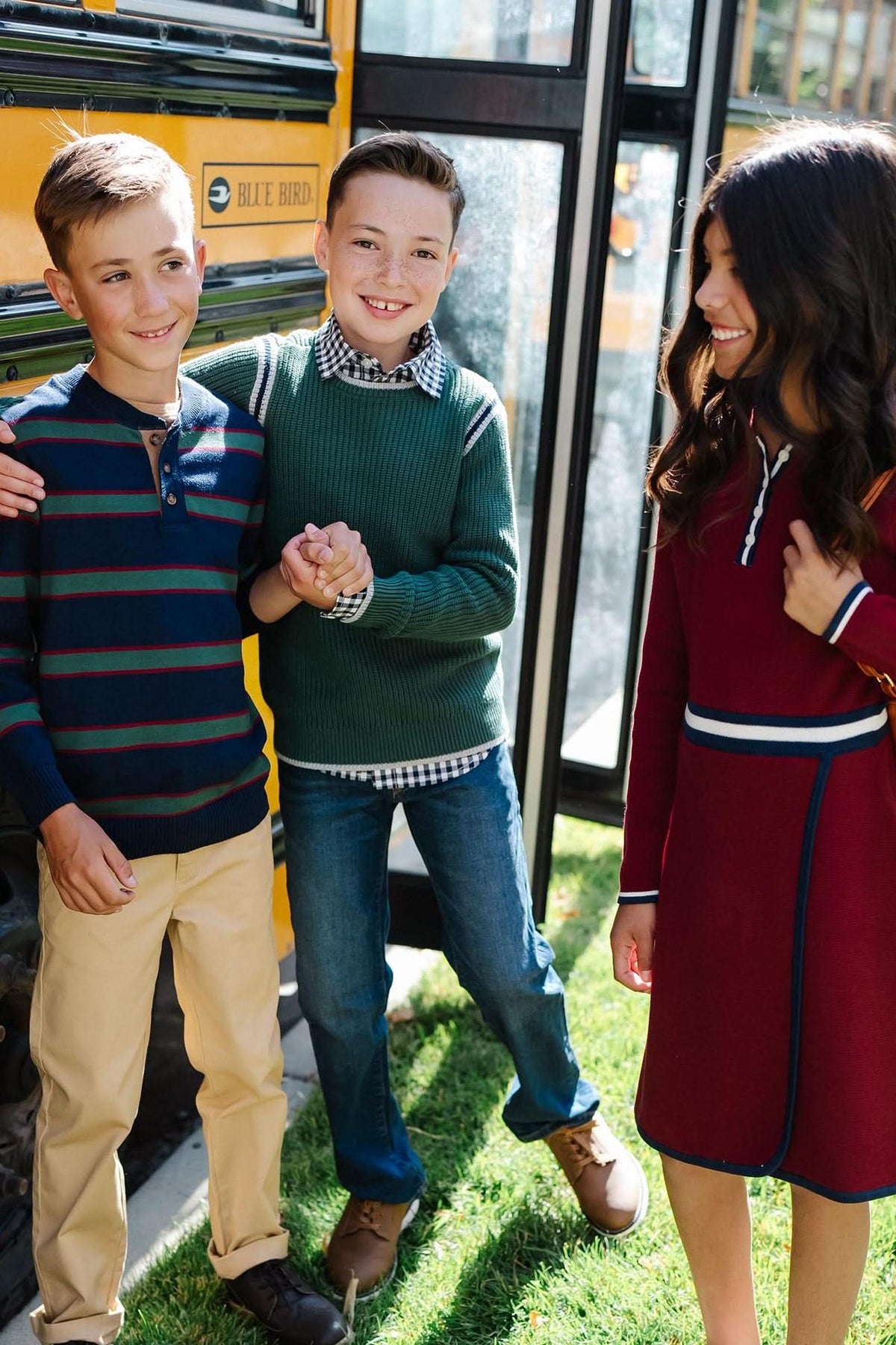 Three children stand by a yellow school bus, wearing colorful sweaters and a dress, smiling and interacting in a grassy.