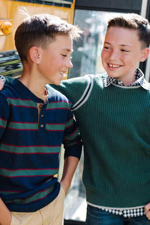 Two boys are smiling and standing together, wearing colorful sweaters, with a school bus in the background.