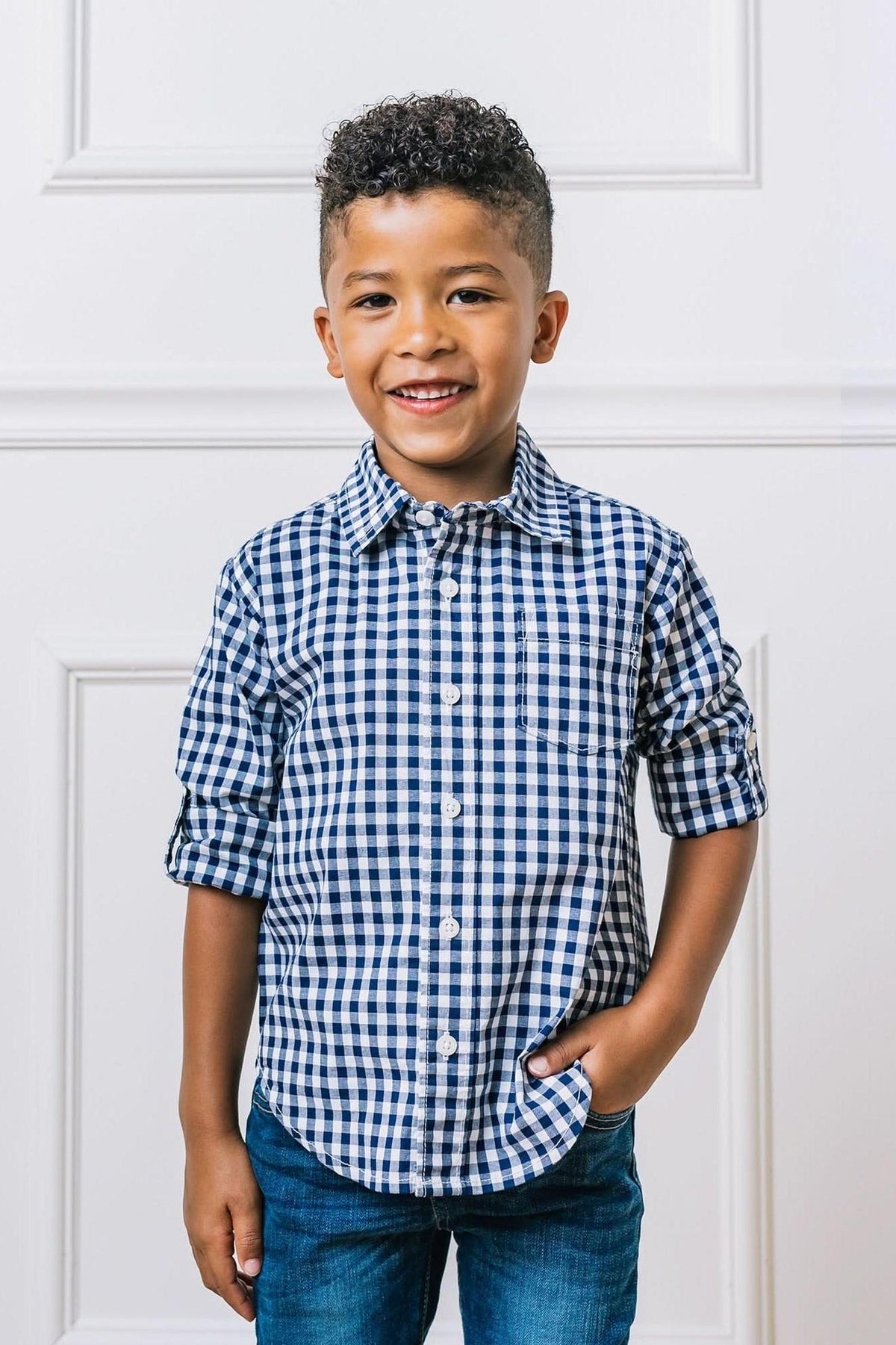 A smiling child models a navy gingham button-down shirt with rolled sleeves, paired with blue jeans against a white background.